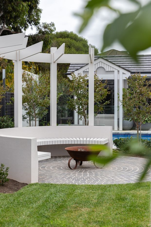 A landscaped outdoor courtyard beside a white rendered garden structure and pergola. The space features curved built-in white seating, a circular grey brick-paved terrace, a sculptural rust-coloured fire bowl, clipped planting, and a blue-edged swimming pool. The background building has white vertical detailing, dark grey shingle-like roofing, and glazed openings.