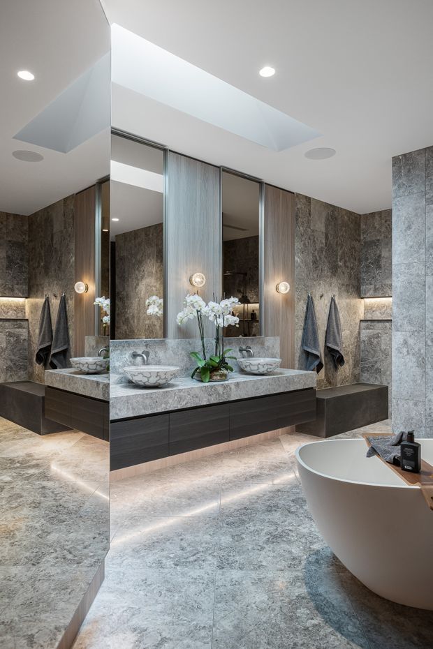 Luxurious contemporary bathroom featuring a freestanding white bathtub, double floating dark-wood vanity with vessel basins, large mirrors, grey stone wall and floor finishes, integrated warm lighting, and a recessed ceiling detail. Towels and orchids provide subtle decorative accents.