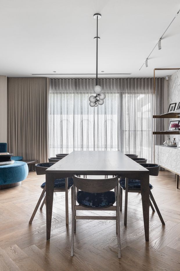 A refined dining room with a large dark timber dining table surrounded by upholstered chairs, set against full-height sheer curtains and heavy grey drapery. The room includes a sculptural clustered pendant light, pale herringbone timber flooring, a deep blue lounge chair at left, and built-in shelving with marble-patterned backing on the right. The interior is bright, contemporary, and carefully composed.