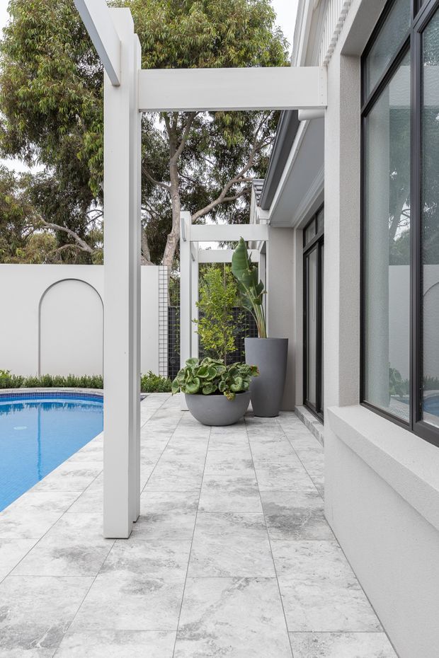 A covered outdoor terrace runs alongside a contemporary white-rendered house, with black-framed glazing, white structural posts and beams, large pale stone-look tiled paving, and a swimming pool visible to the left. Potted plants soften the minimal architectural setting, with mature trees beyond the courtyard wall.