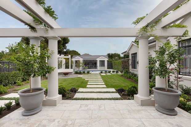 A formal landscaped courtyard viewed from a covered pergola terrace, with white classical columns and beams supporting climbing greenery. Large potted citrus trees frame a pale stone-paved terrace, while a straight stepping-stone path crosses a manicured lawn toward a small garden pavilion. White and grey residential buildings surround the garden.