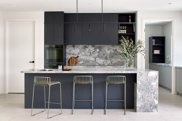 Contemporary kitchen with a large dark island, three bar stools, integrated appliances, extensive dark cabinetry, and a dramatic veined stone splashback and waterfall island end. A white door and open pantry or utility area are visible beyond the kitchen.