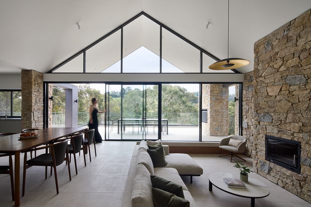 Open-plan living and dining area with a dramatic glazed gable wall and high pitched ceiling. A long timber dining table occupies the left side, while a light neutral sectional sofa and round coffee table face a built-in fireplace set into a prominent rough-stone feature wall. Full-height glazing opens toward a leafy outdoor view and balcony, creating a bright, nature-connected interior.