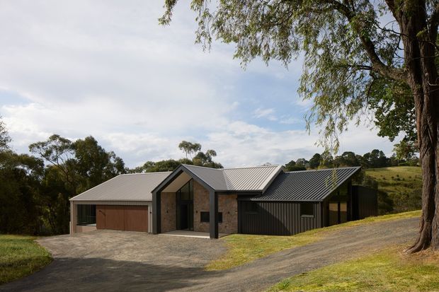 A low-profile contemporary rural house viewed from the driveway, with multiple intersecting pitched metal roofs, dark timber or stained-cladding garage doors, exposed masonry walls, and large glazed openings. The home sits within a gently sloping, tree-lined landscape with a gravel driveway and open pasture beyond.
