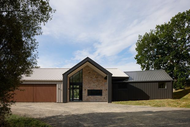 Front exterior view of a low-profile contemporary house with a prominent dark-framed gabled entry volume. The facade combines vertical dark cladding, a warm timber garage door, a light corrugated metal roof, and a central natural stone feature wall with tall glazed entry doors and a dark horizontal opening. The home sits along a gravel driveway and is framed by mature trees and lawn.