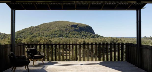 A covered timber deck or terrace framed by dark vertical slatted balustrades and exposed roof structure, furnished with two lounge chairs and overlooking a heavily wooded landscape and distinctive rounded mountain escarpment. The open-sided outdoor space has strong indoor-outdoor character, clean modern detailing, and expansive views.
