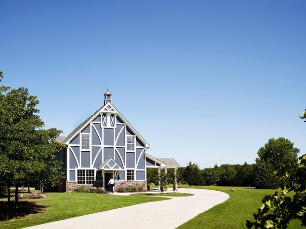 A large barn-inspired building sits in a manicured rural landscape under a clear blue sky. The facade features dark grey panels with prominent white timber-style framing, a steep gabled roof, a central cupola, stonework at the base, and a covered side veranda or walkway. A curved pale driveway approaches the entrance through broad lawns and mature trees.