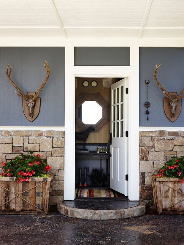 A symmetrical farmhouse-style exterior entry with a white-framed open door, blue-grey wall panels, exposed natural stone masonry, and matching timber planter boxes. Decorative antler mounts flank the doorway, while the entry vestibule beyond contains a small console, octagonal mirror, bench, and striped mat.