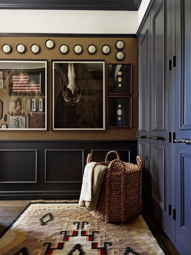 A richly detailed, moody entry hall with deep navy-black raised-panel wall panelling and matching built-in cabinetry, a warm patterned wallcovering, framed artwork, and a display of small decorative plates. A large woven basket with a folded throw sits on a geometric patterned rug over timber flooring. The space has a refined, eclectic heritage-inspired character with strong contrast and layered texture.