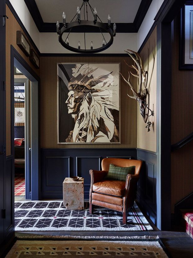 A richly detailed, narrow interior sitting area or hallway with dark navy/black wainscoted panelling, patterned brown wallcovering, and deep contrasting trim. A cognac leather wingback chair sits beside a small stone side table, beneath a large black circular chandelier. The room features a framed illustrated portrait, sculptural wall-mounted antlers, patterned black-and-white carpeting, and views into adjacent rooms through open doorways. The overall aesthetic is moody, refined, eclectic, and traditional with strong lodge-inspired character.