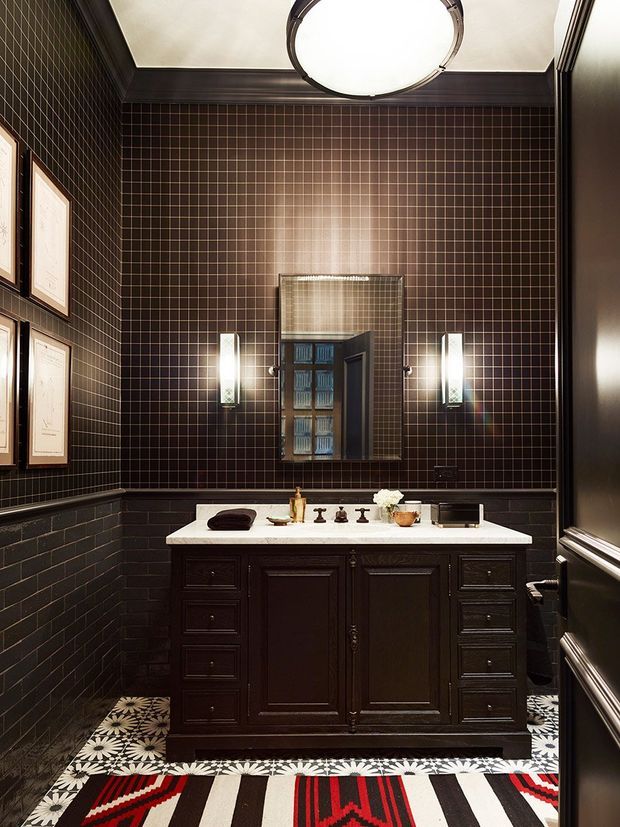 A dramatic, compact bathroom or powder room with dark brown small-format tiled walls, matching dark lower wall tiles, and a traditional dark timber vanity with a white stone benchtop and double basins. A rectangular mirror is centered above the vanity, flanked by vertical wall sconces. Framed artwork lines the side wall, while patterned black, white, and red floor coverings add a bold graphic accent. The room has dark decorative trim and a large circular ceiling light.