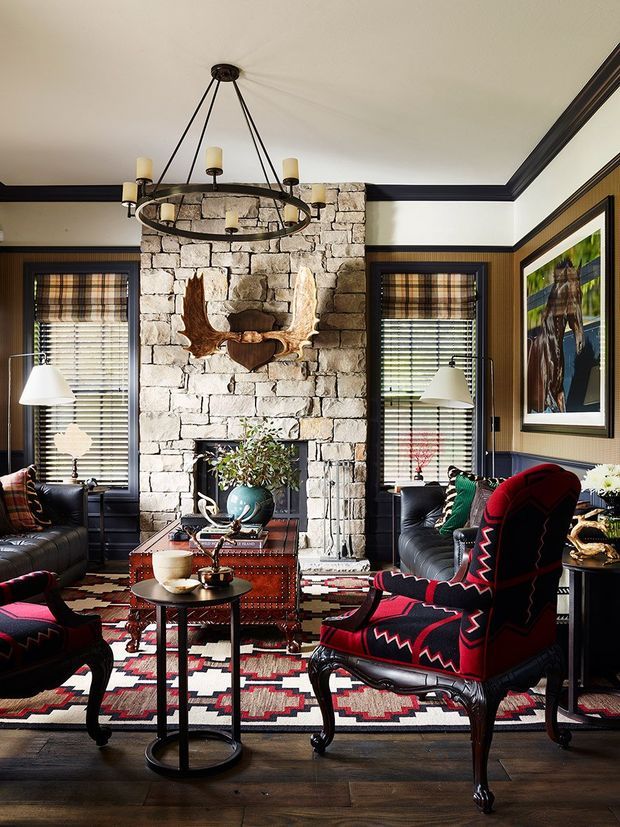 A richly layered living room centred on a substantial rough-cut stone fireplace with antler decor. Dark timber wall panelling and trim frame tall windows with patterned Roman blinds, while leather sofas, a vivid red-and-black patterned armchair, a patterned area rug, an antique-style red chest, and mixed metal and wood tables create a warm, eclectic lodge-inspired interior. A large circular hanging chandelier is suspended from the ceiling.
