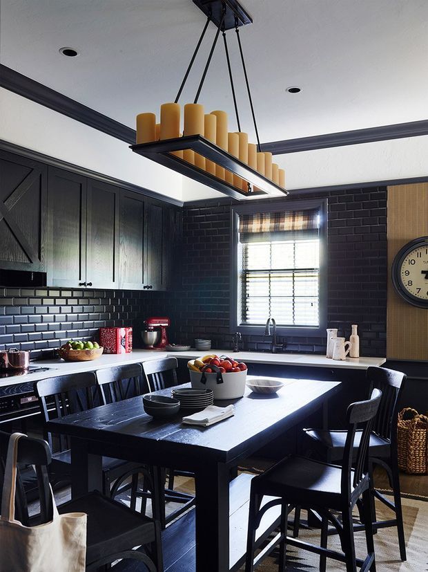 A moody, refined kitchen and dining area with dark charcoal cabinetry, black brick backsplash, and a large black dining island surrounded by chairs. A linear pendant light with warm cylindrical shades hangs above the island, while a window provides natural light. The room includes stainless kitchen appliances, open shelving, a wall clock, woven basket storage, and warm timber accents.