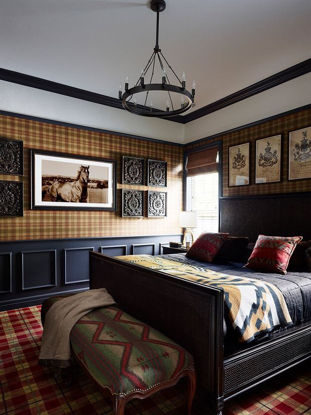 A richly decorated bedroom with dark navy-black wall panelling and trim, red and olive tartan wallpaper and carpet, a substantial dark wood or painted bed, patterned bedding and pillows, and a matching upholstered bench at the foot of the bed. Framed equestrian artwork and decorative panels line the walls, while a large circular pendant chandelier hangs from the ceiling. The room has a dramatic, traditional, tailored character with strong pattern and contrast.