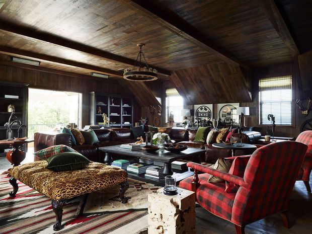 A richly furnished rustic lounge or living room with dark timber wall and ceiling panelling, exposed heavy beams, built-in shelving, leather sofas, a large black coffee table, a patterned chaise longue, and bold red plaid armchairs. Natural light enters through large windows with blinds, while a circular chandelier and layered rugs add character. The room has a warm, eclectic lodge-like atmosphere with substantial timber detailing and vintage-inspired furnishings.