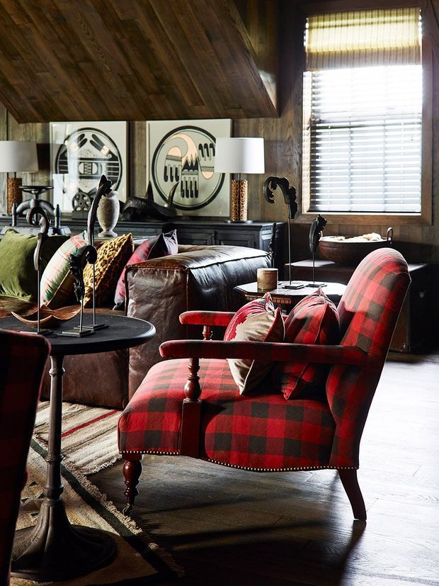 A richly layered living room with a strong lodge-inspired character. The room features a steeply sloped timber-clad ceiling and matching dark timber wall finishes, a large window with layered blinds, a dark leather sofa, a striking red-and-black tartan upholstered armchair, patterned cushions, a dark round pedestal table, lamps, framed artwork, and traditional decorative objects. The palette is warm, earthy, and dramatic, with abundant wood, leather, tartan, and vintage-style furnishings.