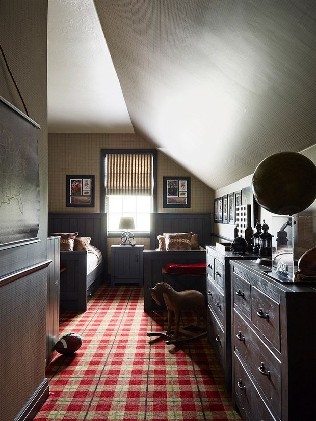 A narrow, richly detailed attic-style bedroom or retreat with a steep sloped ceiling, central window with blinds, twin built-in daybeds or single beds, dark wood wall panelling and cabinetry, framed artwork, and a bold red-and-green plaid carpet. A dresser with decorative objects lines the right side, while a rocking horse and sports ball add a playful character. The room has a warm, traditional, slightly masculine atmosphere with strong natural light.