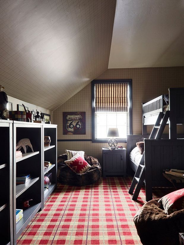 A compact, cozy bedroom with a steep sloped ceiling, dark built-in shelving and bunk-style bed, a central window with striped Roman blind, plaid carpet, and rustic furnishings including fur-covered seating and red patterned cushions. The room has a warm, cabin-like atmosphere with strong red, cream, brown, and charcoal tones.