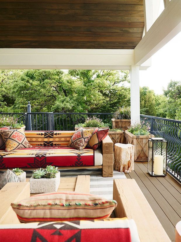 A covered outdoor terrace or balcony furnished as a relaxed lounge, with a timber-lined ceiling, white structural beams and posts, dark metal railing, timber decking, and built-in or substantial timber seating upholstered in bold red, cream, and black geometric textiles. Planters with succulents and greenery surround the seating, and a freestanding lantern adds a warm decorative element. The terrace overlooks dense mature trees.