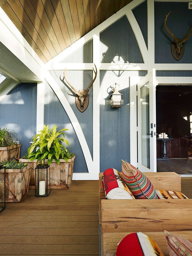 A bright, eclectic indoor-outdoor sitting area with a timber-lined sloping ceiling, dark blue and white geometric wall panelling, timber decking, rustic planter boxes, and a natural wood sofa with colourful patterned cushions. Decorative deer antlers and a pendant lantern add a lodge-inspired character. An open glazed door leads into a darker adjoining interior space.