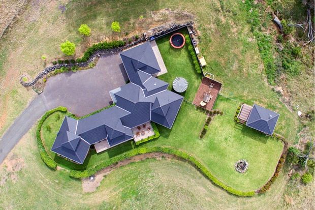 Aerial view of a large detached residence set within extensive landscaped grounds. The complex has multiple steep pitched, dark tiled roof sections, connected wings, paved driveway and paths, manicured hedges, a separate small outbuilding, outdoor seating area, circular trampoline or play feature, and a firepit area.