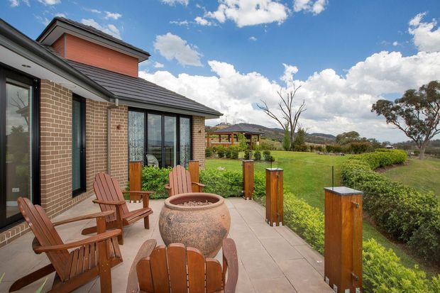 A residential outdoor terrace beside a brick house, furnished with timber Adirondack-style chairs around a large round fire bowl. The paved terrace has timber-and-metal balustrade posts overlooking a broad landscaped lawn and rural-looking landscape. The house features dark tiled roof planes, reddish-brown brick walls, large glazed doors and windows, and a raised painted upper wall section.