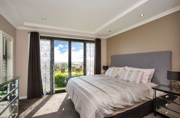 Bright, refined bedroom with a large upholstered grey headboard, neatly made king-size bed, mirrored bedside tables, layered curtains, and floor-to-ceiling glazed doors opening to a garden and distant suburban view. The room features a tray ceiling with decorative moulding and recessed downlights, neutral taupe walls, and soft grey-beige furnishings.