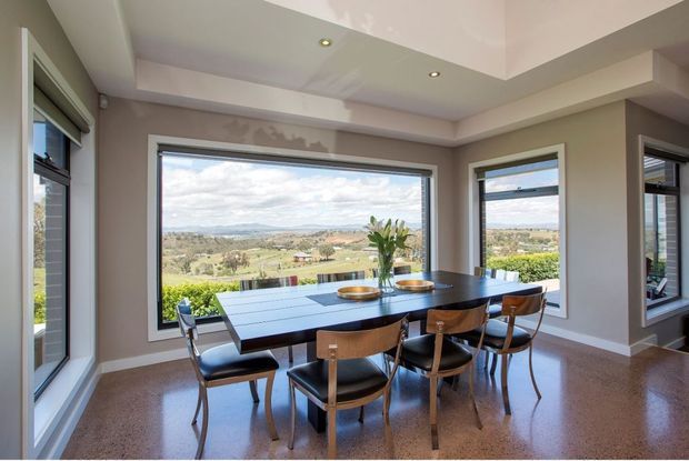 Bright dining area with a rectangular dark timber dining table and matching chairs, surrounded by large windows overlooking a rural landscape. The room has warm neutral walls, polished concrete-style flooring, recessed lighting, and a stepped tray ceiling.