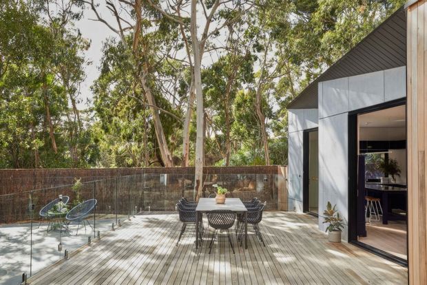 Contemporary timber-decked outdoor entertaining area beside a modern pavilion, surrounded by mature eucalyptus trees. The deck includes an outdoor dining table with woven chairs and a small seating area, enclosed by a timber privacy fence. Large black-framed glazed doors open into an interior kitchen or living space, while the pavilion features light vertical timber cladding, dark metal soffits, and pale grey geometric wall panels.
