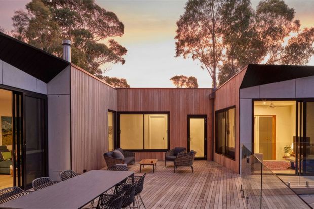 A contemporary single-storey courtyard residence arranged around a spacious timber deck. The image shows angular pavilion-like wings with dark metal roof planes, vertical timber wall cladding, large dark-framed glazed openings, and a central outdoor dining and lounge area. Warm interior lighting and a sunset sky create a relaxed, inviting atmosphere, with mature eucalyptus trees visible beyond the courtyard.