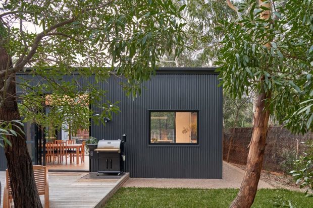 Exterior view of a compact dark-clad backyard building or extension, set among established eucalyptus trees. The vertically ribbed dark facade has a single window and opens onto a timber deck with an outdoor dining area and barbecue. Gravel edging, lawn, and natural vegetation surround the structure.