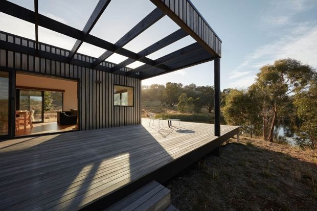 A contemporary elevated timber-clad building viewed from a broad timber deck and partially covered outdoor terrace. Dark vertical cladding, exposed roof beams, and slender black structural framing define the open pavilion-like form. Through the glazing, a simple dining area and living space are visible. The deck overlooks native trees and a small body of water, creating a quiet bushland setting.