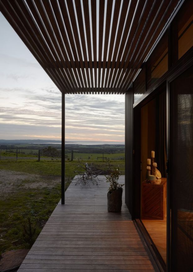 A covered timber deck or verandah extends along the side of a dark-clad building, with a slatted timber pergola overhead. Large glazed sliding doors open to a warmly lit interior with timber joinery and decorative objects. Beyond the deck is an expansive rural landscape and distant horizon under a cloudy sunset sky.