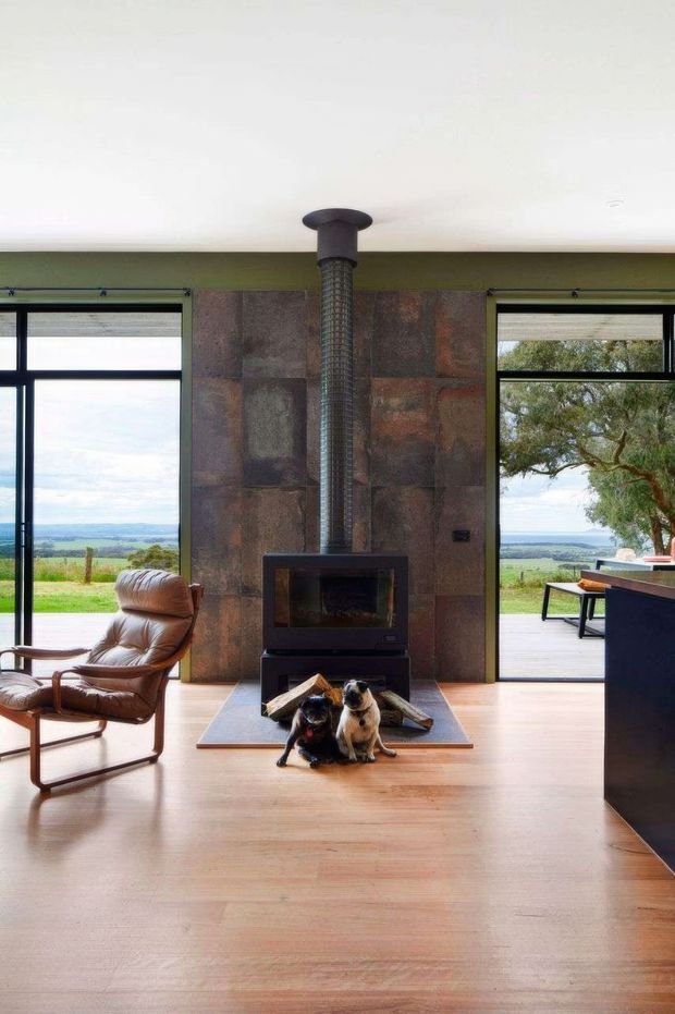 A contemporary living room centered on a freestanding black fireplace with a tall flue set against a large rust-toned stone feature wall. Full-height glazed openings on both sides provide views to a rural landscape and outdoor deck. The room has a pale timber floor, white ceiling, dark window frames, and a vintage-style leather lounge chair. Two dogs sit on the hearth in front of the fireplace.