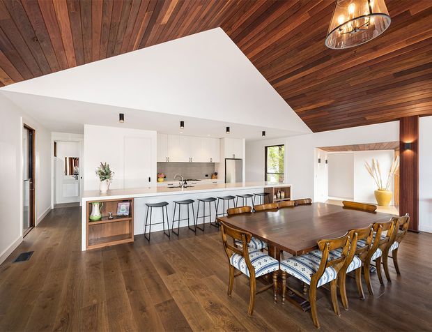 Open-plan kitchen and dining area with a large white island, integrated white cabinetry, dark timber flooring, and a dramatic warm timber-lined ceiling. A substantial timber dining table with upholstered dining chairs occupies the foreground, while pendant and recessed lighting create a bright, welcoming contemporary interior.
