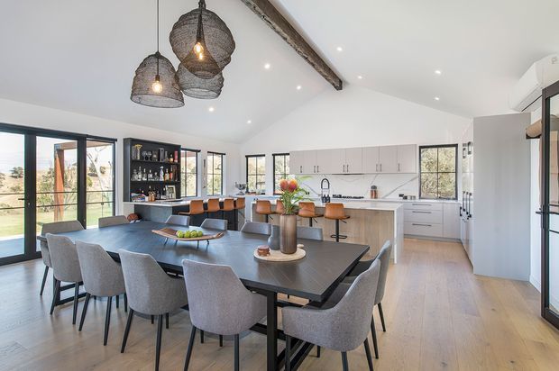 Bright open-plan kitchen and dining area with a large dark dining table surrounded by upholstered chairs, a white island with bar seating, pale cabinetry, extensive black-framed glazing, and a high pitched ceiling with an exposed timber beam. Woven pendant lights and warm timber flooring add texture and character. A living or outdoor area is visible beyond the glazing.