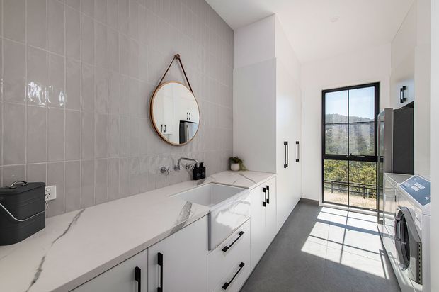 Bright contemporary laundry room with a long white stone benchtop, integrated utility sink, extensive white cabinetry, grey tiled splashback wall, black hardware, front-loading washing machine, and a large black-framed window overlooking greenery. A round hanging mirror and small decorative planter add detail.