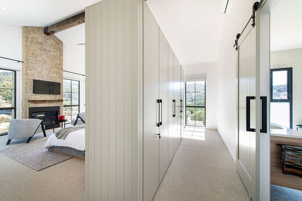 Bright contemporary bedroom suite with a long wall of pale built-in wardrobes and mirrored panels forming a corridor-like dressing area. The bedroom includes a low bed, upholstered lounge chair, fireplace with stone surround, timber beam, large windows, and soft neutral carpeting. A bathroom or vanity area is partially visible through the opening on the right.