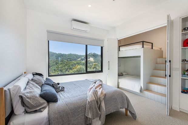 Bright compact bedroom with a double bed, grey bedding, built-in bunk sleeping alcove, and integrated open shelving. A large window overlooks a green, hilly landscape, while white walls, light carpet, and simple recessed lighting create a clean, contemporary feel.
