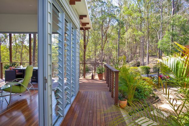 A sunlit timber deck and covered outdoor walkway beside a glazed, louvred residential facade. The deck has timber flooring, dark timber and metal balustrades, potted plants, and views into a native woodland garden. Through the glazing, a small interior seating area is visible. The architecture features horizontal louvres, light-coloured cladding or wall panels, and exposed red-brown structural elements.