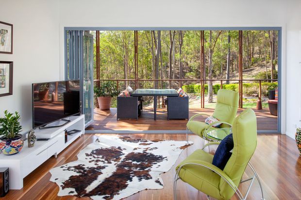 Bright living room opening onto a timber deck through expansive floor-to-ceiling glazing. The room features warm timber flooring, a white low-profile media cabinet, lime-green lounge chairs, a cowhide rug, and a compact outdoor dining setting beyond the glazing. The surrounding wooded landscape is visible outside.