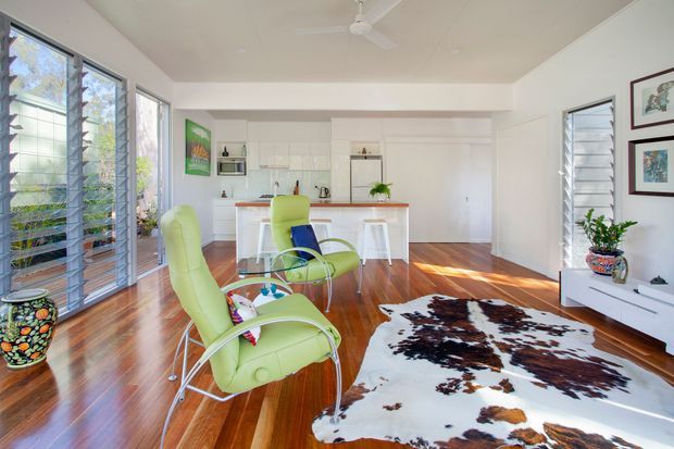 Bright open-plan living and kitchen interior with polished timber flooring, white cabinetry, extensive louvred glazing, lime-green mid-century-style lounge chairs, and a cowhide rug. A compact kitchen island with stools sits at the rear, while a white media console and framed artwork occupy the right wall. A ceiling fan and abundant daylight create an airy, relaxed atmosphere.