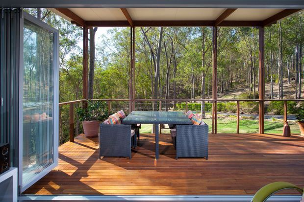A covered timber deck or enclosed outdoor dining area opening onto a wooded landscape. The space features warm timber decking, slender timber posts and beams, extensive floor-to-ceiling glazing, a glass-topped outdoor dining table with woven chairs, and colourful cushions. The setting feels light, natural, and connected to the surrounding bush.