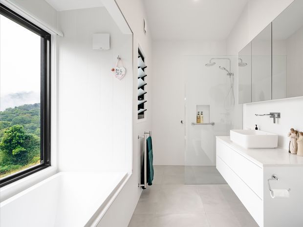 Bright contemporary bathroom with a freestanding-style white bathtub beside a large black-framed window, a frameless glass walk-in shower, wall-mounted white vanity with vessel basin, mirrored cabinetry, and pale grey floor tiles. The room has a clean, minimalist fit-out with abundant natural light and a leafy landscape visible outside.