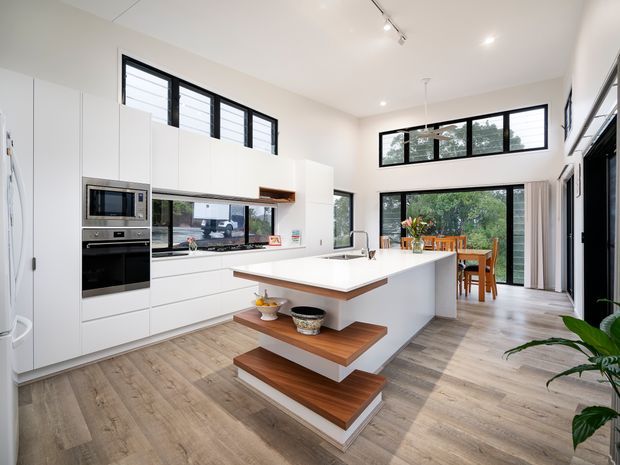 Bright contemporary open-plan kitchen and dining area with white floor-to-ceiling cabinetry, integrated stainless-steel appliances, a large white island with timber trim and floating timber display shelves, and light timber-look flooring. High-level horizontal windows, skylights or track lighting, and expansive glazing create an airy connection to the outdoor garden and dining area.