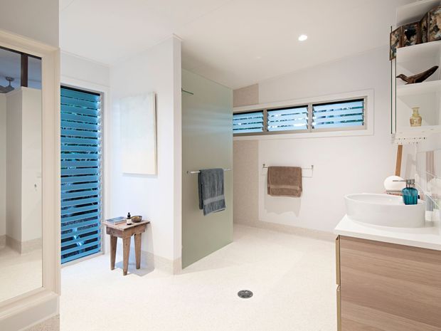 Bright contemporary bathroom with a walk-in wet-area layout, frameless glass shower screen, freestanding white vessel basin on a timber-look vanity, and horizontal louvred windows providing natural ventilation. White walls and floor create an airy, clean interior, with open shelving and towel rails adding practical storage and detail.
