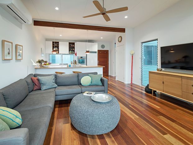 Bright open-plan living room with a large grey sectional sofa and round upholstered ottoman in the foreground, facing a timber media console and wall-mounted television. The room opens to a compact kitchen with white cabinetry and a peninsula, while polished timber flooring, a timber ceiling beam, recessed lighting, ceiling fan, and a glazed exterior door create a warm contemporary interior.