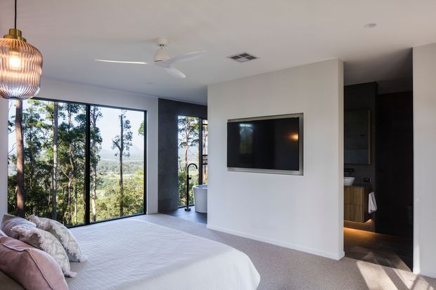 Bright contemporary bedroom with a large upholstered bed, expansive floor-to-ceiling glazing overlooking trees, a wall-mounted television, recessed lighting, ceiling fan, and a dark ensuite or bathroom area visible through an opening. The room uses a soft neutral palette with carpeted flooring and a sculptural pendant light.