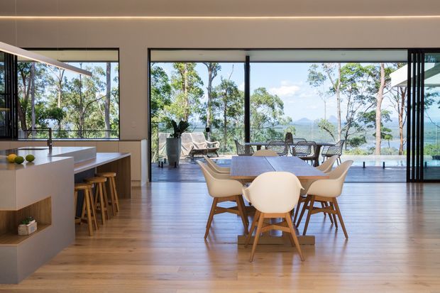 A bright, open-plan contemporary dining and kitchen interior with extensive floor-to-ceiling glazing overlooking a leafy, mountainous landscape. The room features warm timber flooring, a streamlined white kitchen island with timber stools, and a timber dining table surrounded by sculptural cream chairs. A large outdoor terrace is visible beyond the glazing.