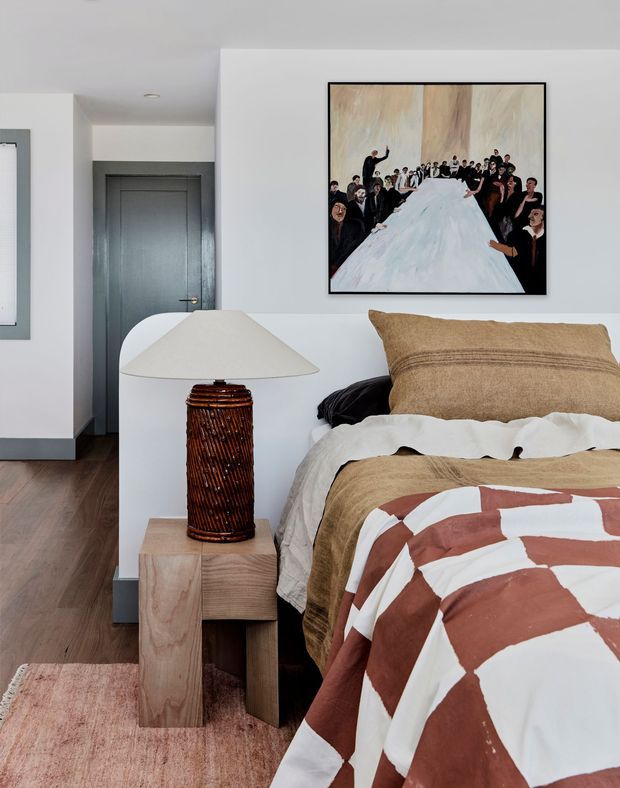 A warmly styled bedroom featuring a bed with layered beige bedding and a rust-and-white geometric quilt, a sculptural wooden bedside table, and a ribbed dark wood table lamp with a broad cream shade. The room has white walls, dark grey interior doors, timber flooring, a muted rug, and a large framed artwork above the bed.